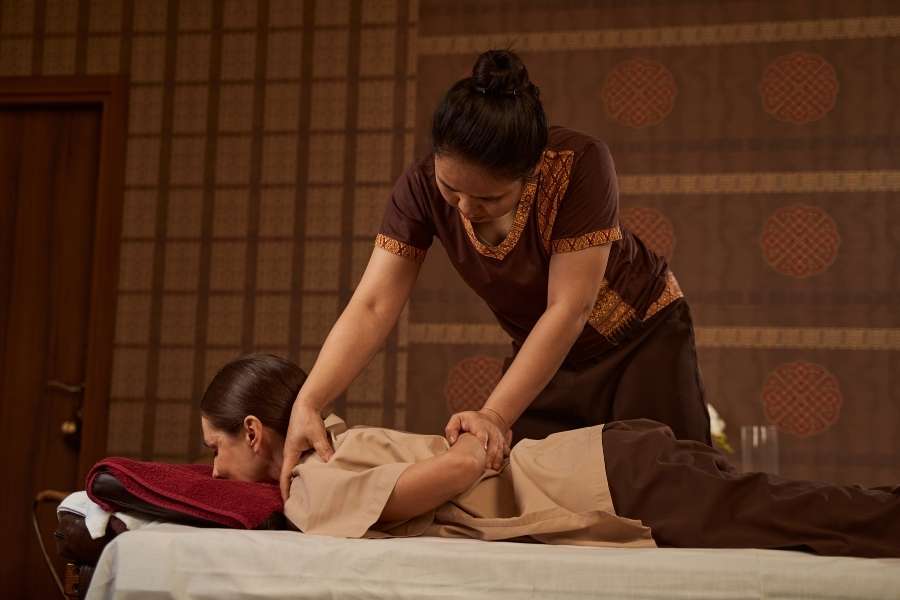 Une masseuse thaïlandaise en tenue traditionnelle effectue une manœuvre d'étirement du dos sur une cliente allongée.