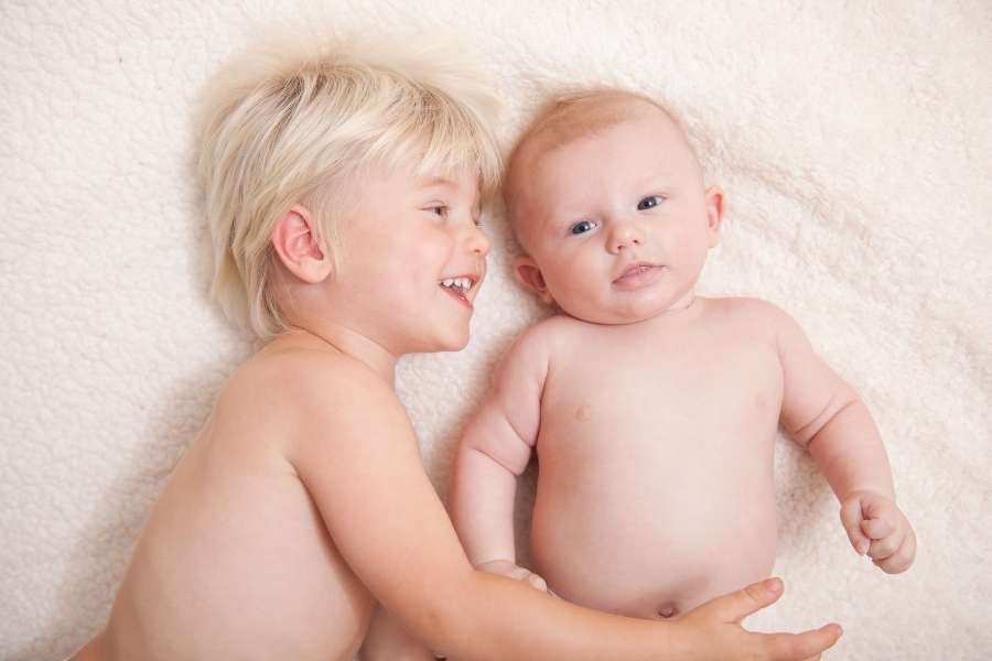 Un jeune enfant blond sourit en regardant un bébé allongé à ses côtés, illustrant la joie de l'arrivée d'un petit frère.